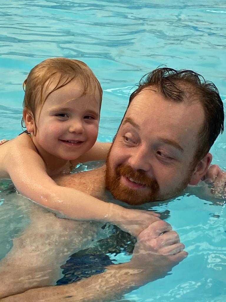 A smiling toddler with their arms wrapped around their dad's shoulders in the pool